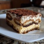A perfect slice of layered tiramisu cheesecake dusted heavily with cocoa powder, served on a white plate.