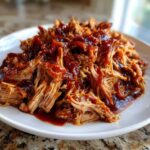 Close-up of tender, shredded Slow cooker pulled pork generously topped with rich, dark BBQ sauce on a white plate.