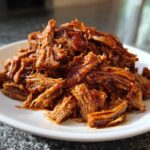 A close-up of perfectly shredded, saucy Slow cooker pulled pork piled high on a white plate.