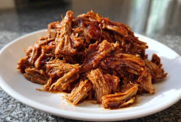 A close-up of perfectly shredded, saucy Slow cooker pulled pork piled high on a white plate.