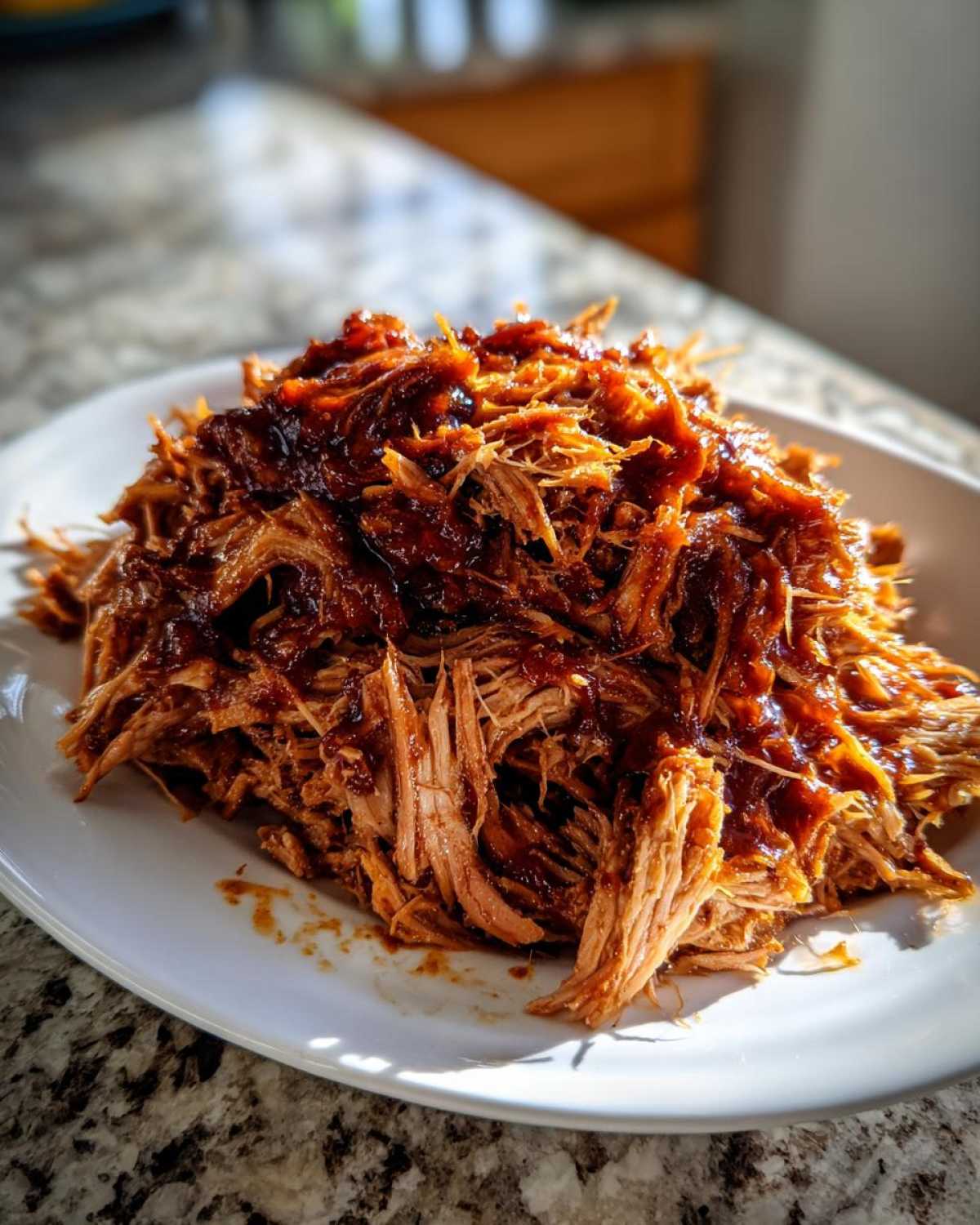 A close-up of shredded, saucy Slow cooker pulled pork piled high on a white plate, glistening under natural light.