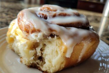 A close-up shot of a soft, fluffy cinnamon roll with a generous swirl of white icing, showing a bite taken out.