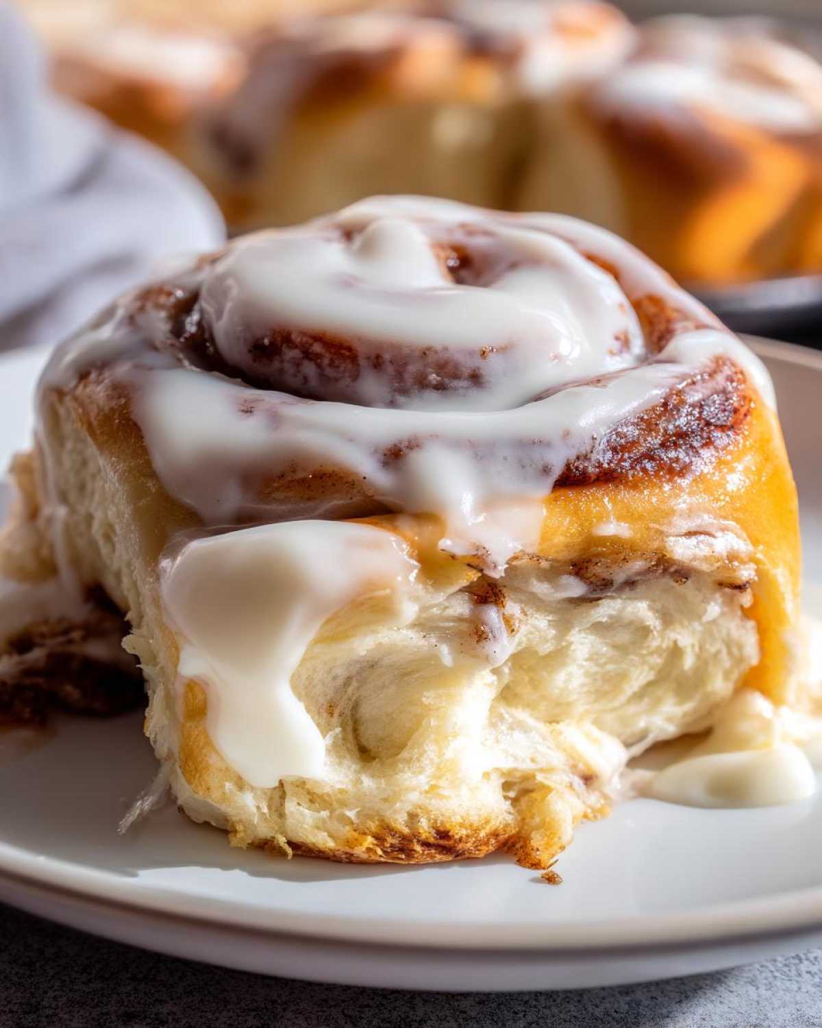 A close-up of a single, fluffy cinnamon roll generously topped with dripping white cream cheese icing.