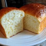 A golden-brown loaf of soft condensed milk bread sliced in half, showing the fluffy white interior.