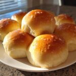 A cluster of golden-brown, soft dinner rolls brushed with butter, sitting on a white plate.