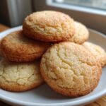 A close-up of several golden brown soft sugar cookies stacked on a white plate, coated in sparkling sugar.