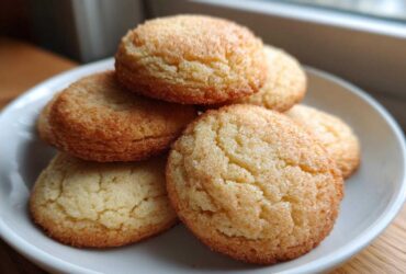 A close-up of several golden brown soft sugar cookies stacked on a white plate, coated in sparkling sugar.