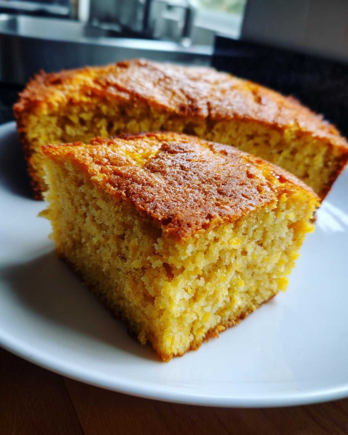 A close-up of a moist slice of golden sourdough cornbread served on a white plate, with the rest of the loaf blurred in the background.