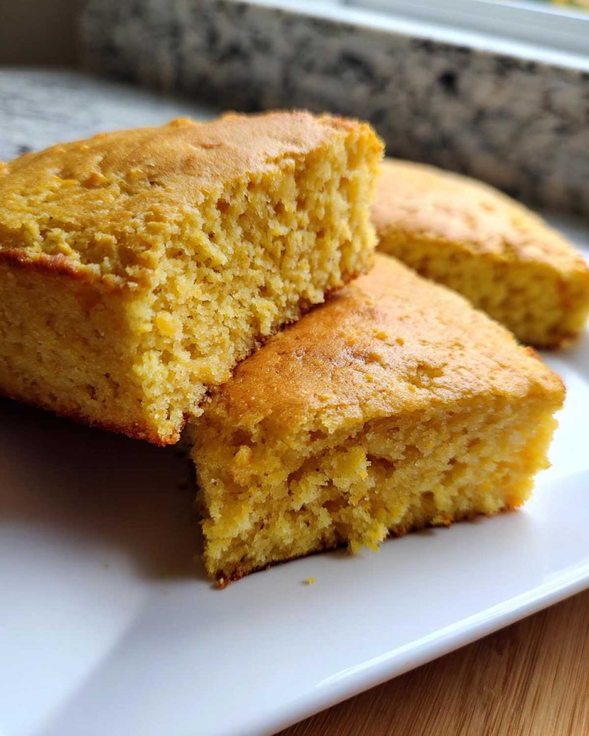 Close-up of three golden squares of moist sourdough cornbread stacked slightly on a white plate.