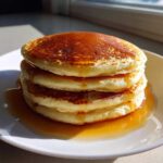 A tall stack of four fluffy sourdough pancakes drizzled generously with golden maple syrup on a white plate.