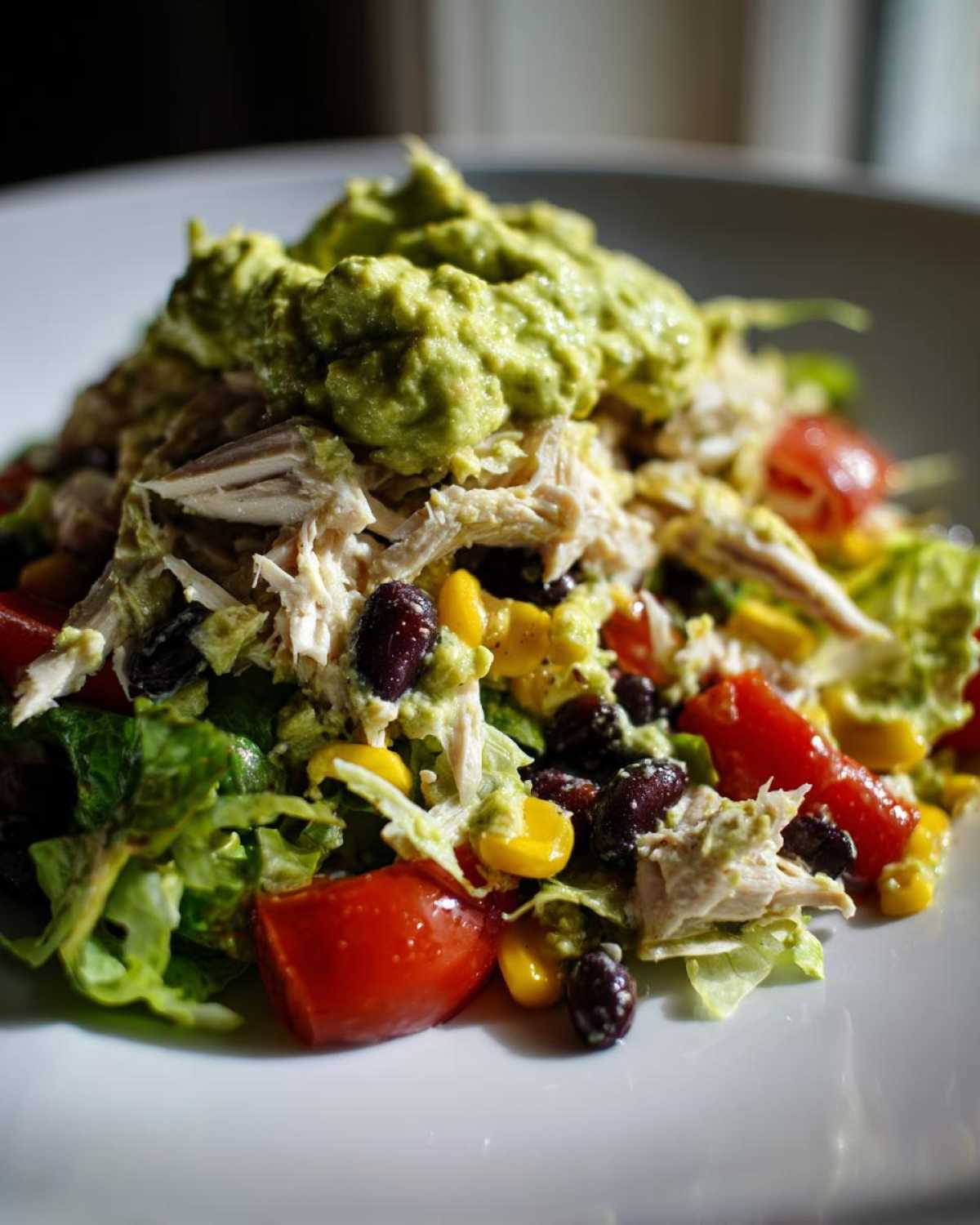 Close-up of southwest chicken salad featuring shredded chicken, black beans, corn, tomatoes, and a dollop of guacamole.