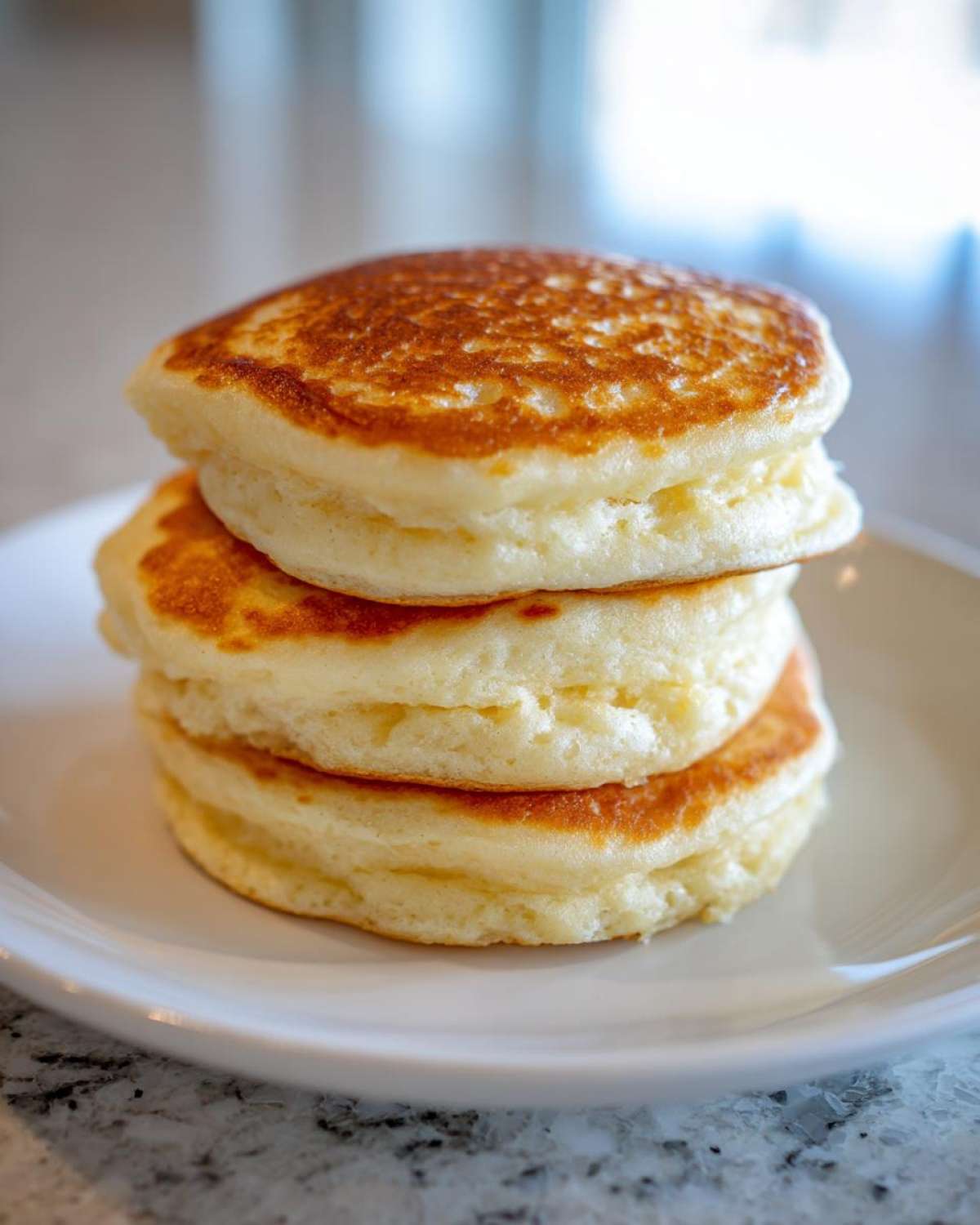 A stack of three incredibly fluffy pancakes with golden-brown tops served on a white plate.