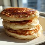 A close-up stack of three thick, fluffy pancakes with perfectly golden brown tops sitting on a white square plate.