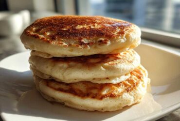 A close-up stack of three thick, fluffy pancakes with perfectly golden brown tops sitting on a white square plate.