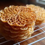 A close-up stack of freshly made, golden brown pizzelle cookies with intricate lace patterns resting on a white cooling rack.