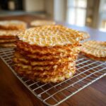A tall stack of golden brown, crisp pizzelle cookies resting on a wire cooling rack.