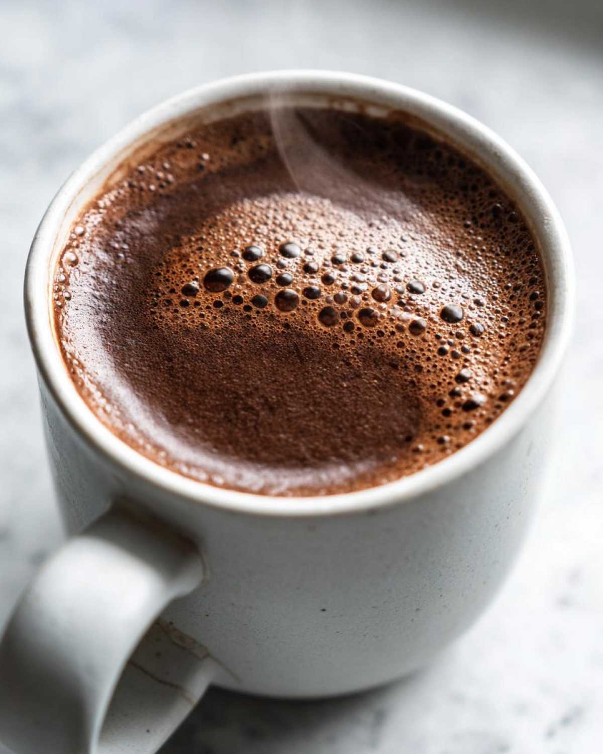 Close-up of a steaming mug filled with rich, dark homemade hot cocoa topped with small chocolate bubbles.