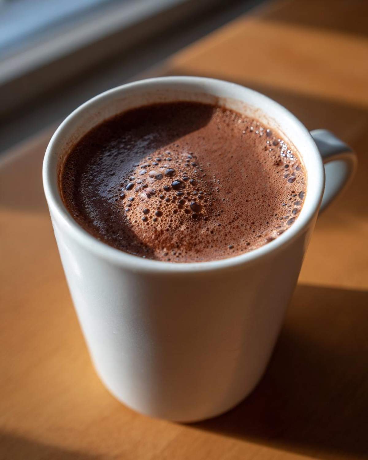 Close-up of a white mug filled with rich, frothy hot cocoa made from homemade hot cocoa mix, sitting on a wooden surface in sunlight.