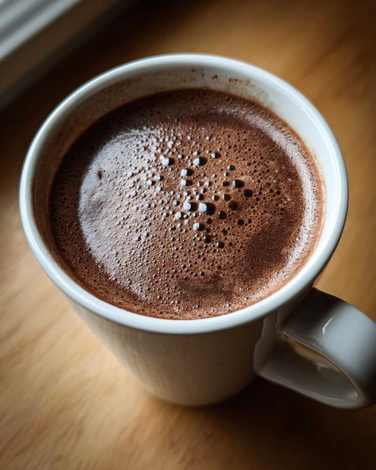 Close-up overhead shot of a white mug filled with rich, foamy hot cocoa mix on a wooden surface.