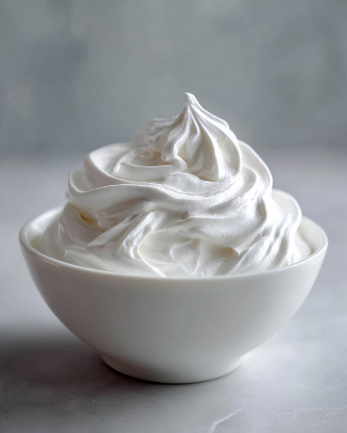 A close-up of stiff, bright white royal icing recipe mixture, piped into a swirl peak in a small white bowl.