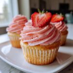 A close-up of a vanilla strawberry cupcake topped with pink frosting and a fresh strawberry slice.