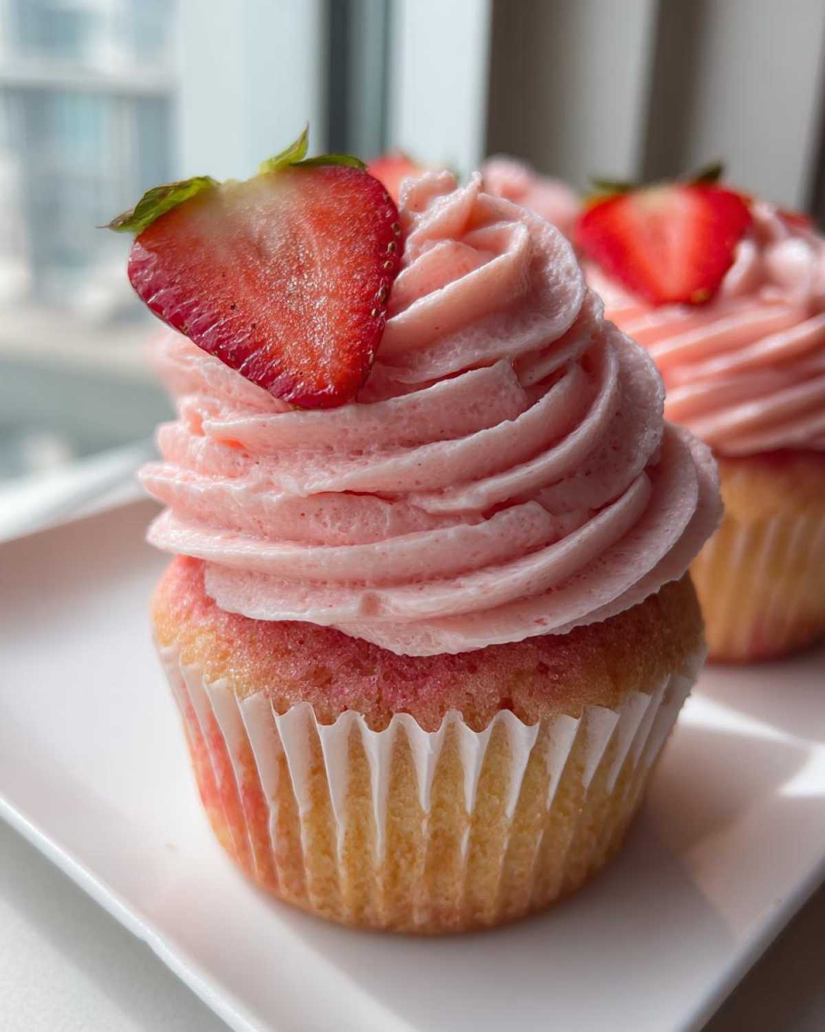 A close-up of a perfectly frosted strawberry cupcake topped with a fresh strawberry slice.