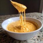 A spoonful of thick, orange beer cheese dip showing amazing cheese pulls over a white bowl.