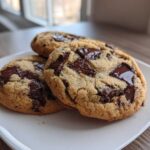 Three freshly baked vegan chocolate chip cookies piled on a white plate with melted chocolate chunks.