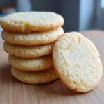A stack of five golden vegan sugar cookies with one leaning against the side on a wooden surface.