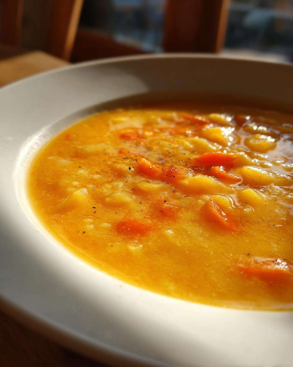 Close-up of a thick, orange cheese soup filled with chunks of potato and carrot, seasoned with pepper, served in a white bowl.