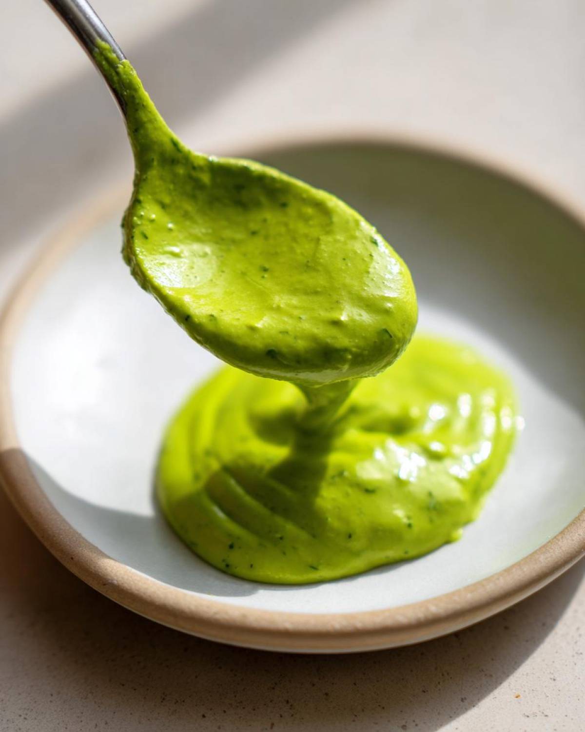 A spoonful of vibrant, bright green Creamy Avocado Cilantro Sauce being drizzled onto a small white plate.