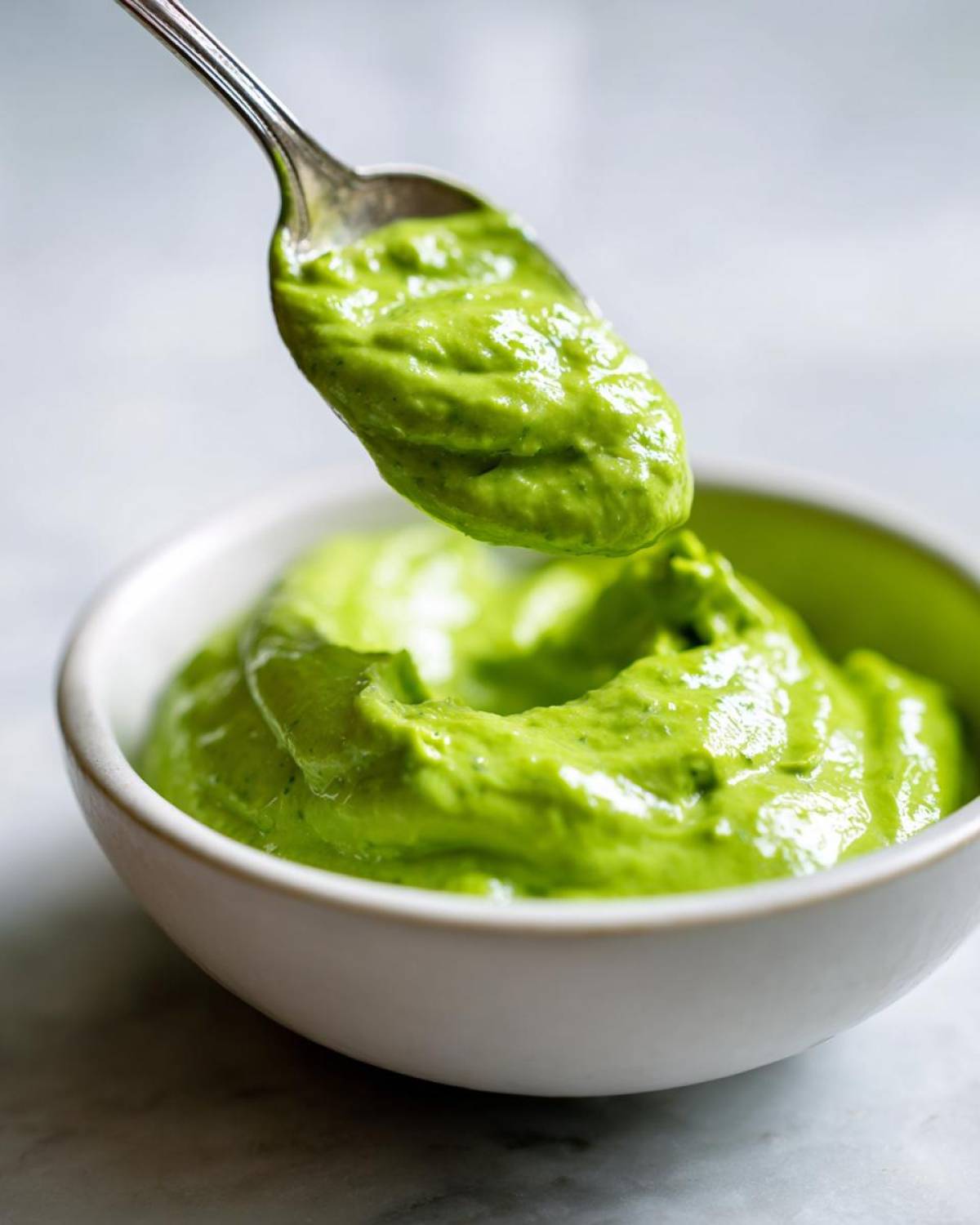 A spoonful of vibrant green Creamy Avocado Cilantro Sauce being lifted from a small white bowl.