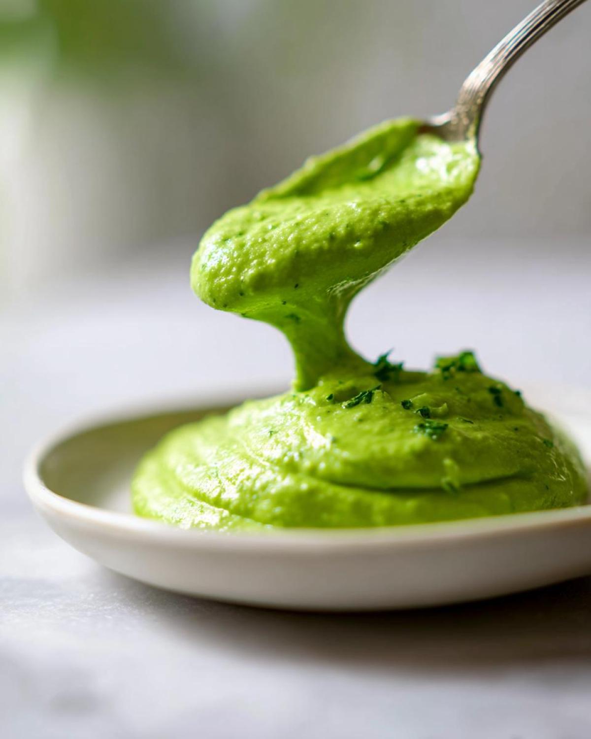 A spoonful of vibrant green, thick Creamy Avocado Cilantro Sauce being lifted from a small white bowl.