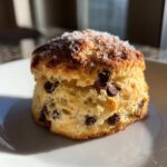 A single, tall Gluten-Free Chocolate Chip Scone topped with coarse sugar crystals, sitting on a white plate.