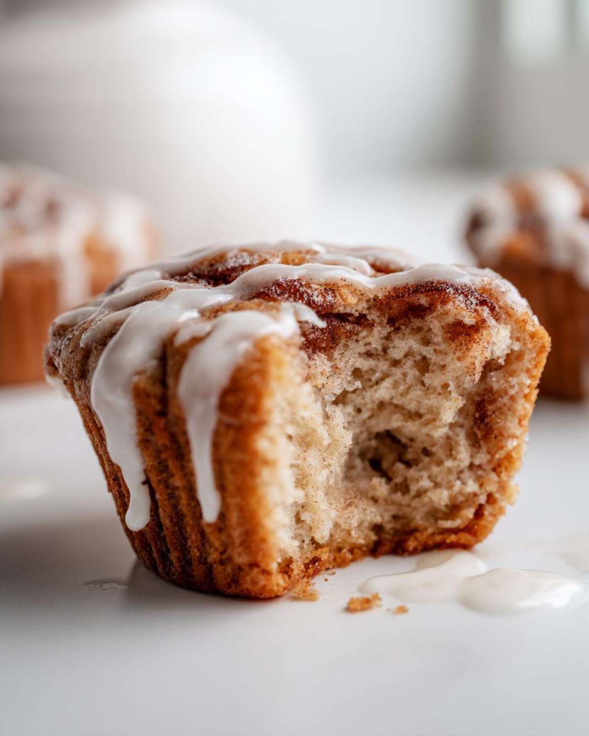 Close-up of a Gluten-Free Cinnamon Roll Muffin with a bite taken out, showing the soft interior and dripping white icing.