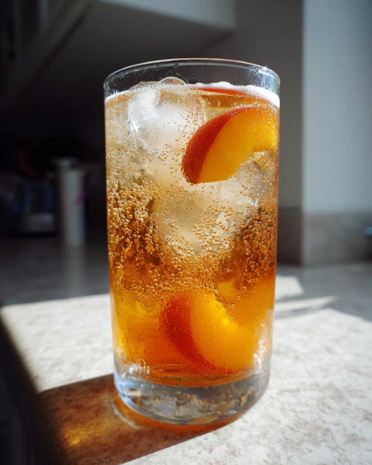 Close-up of a tall glass filled with bubbly Iced Peach Green Tea, ice cubes, and fresh peach slices.