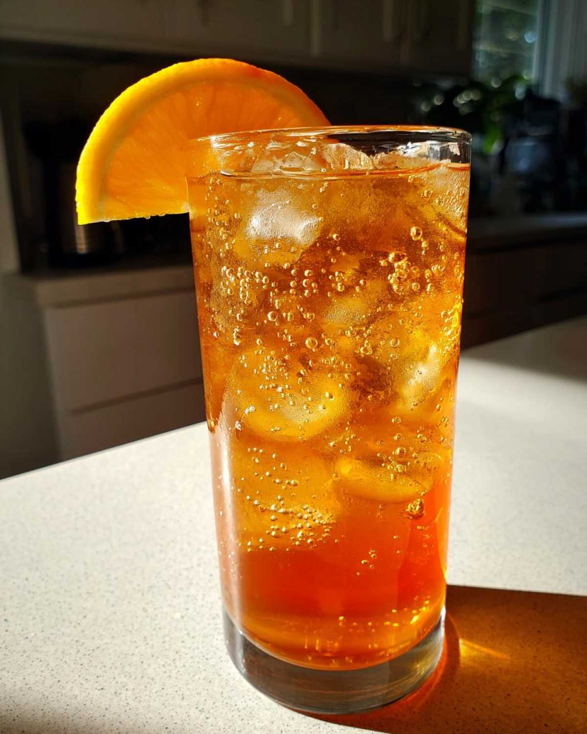 Close-up of a tall glass filled with bubbly, amber Iced Peach Green Tea and ice, garnished with an orange slice.
