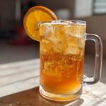 A tall glass mug filled with bubbly Iced Peach Green Tea, ice cubes, and garnished with a bright orange slice.
