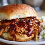 A close-up of a juicy Instant Pot BBQ Pulled Chicken Sandwich piled high with shredded chicken and dripping BBQ sauce, served with coleslaw.