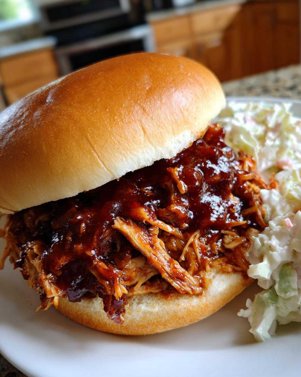 Close-up of a delicious Instant Pot BBQ Pulled Chicken Sandwich piled high with saucy meat on a brioche bun next to coleslaw.