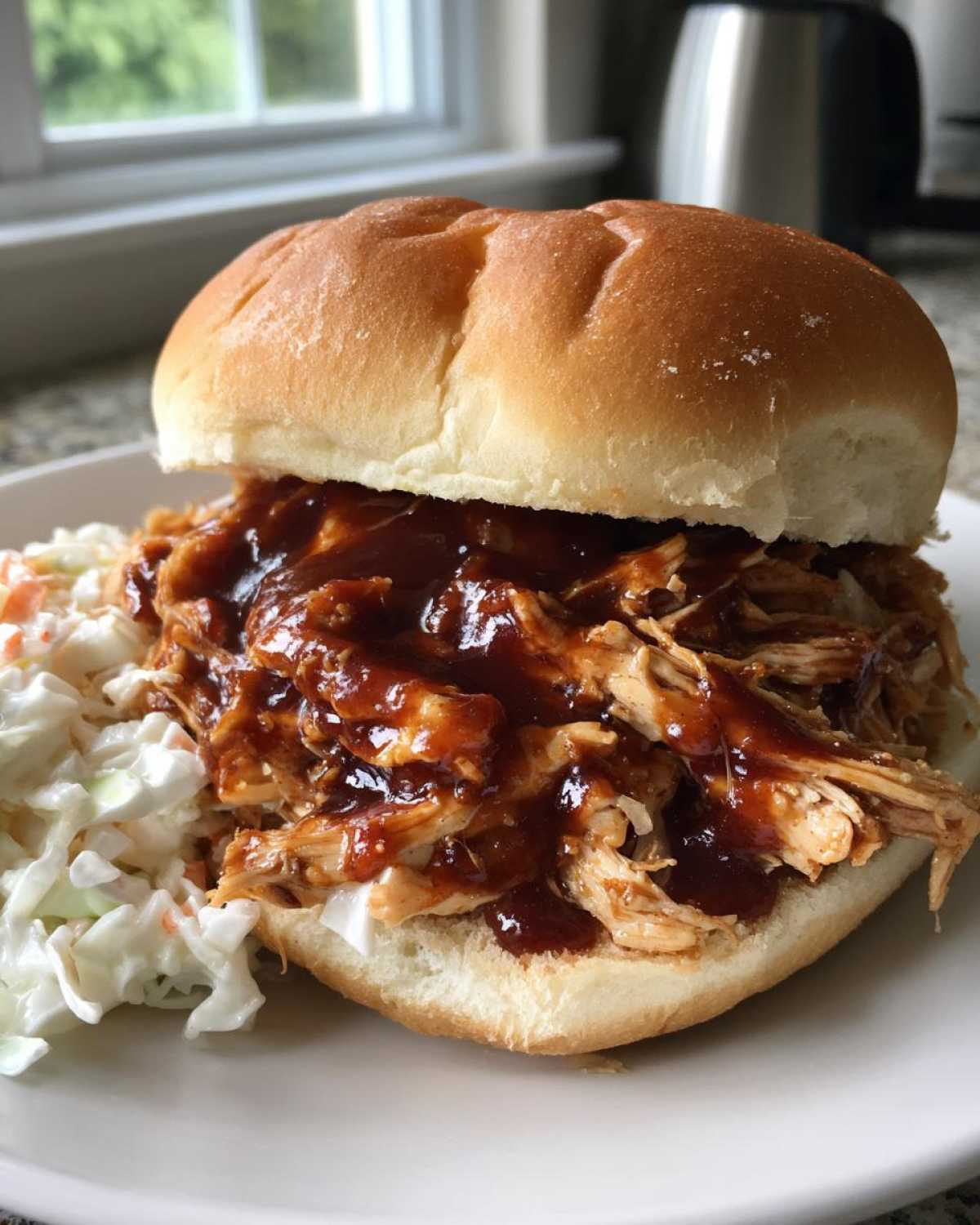 A close-up of an Instant Pot BBQ Pulled Chicken Sandwich piled high with sauce, served next to coleslaw on a white plate.