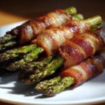 Close-up of perfectly cooked Keto Bacon Wrapped Asparagus Bundles stacked on a white plate.