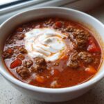 Close-up of a white bowl filled with rich, red Keto Taco Soup with Ground Beef, topped with a swirl of sour cream.