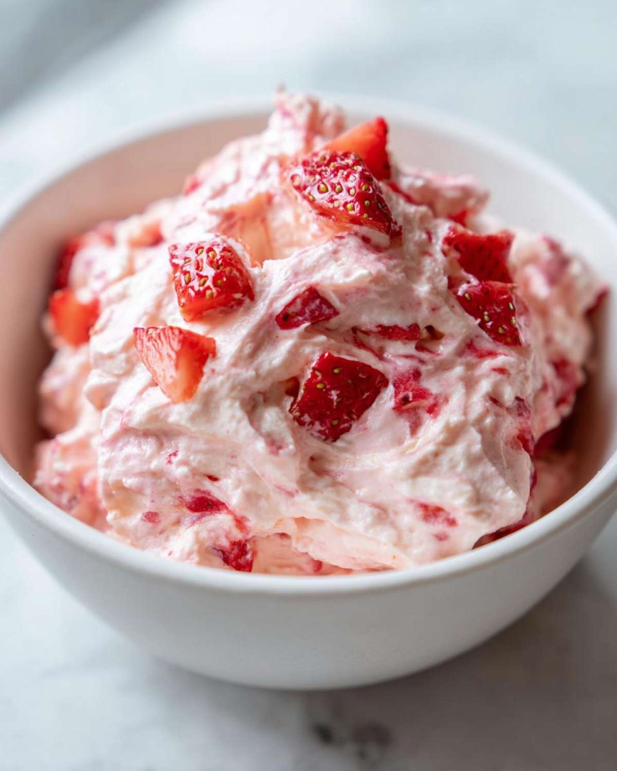 A close-up of creamy, pink No-Bake Strawberry Cheesecake Dip topped with diced fresh strawberries in a white bowl.