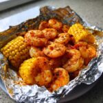 Close-up of cooked shrimp seasoned with Cajun spices next to corn on the cob inside a foil packet.