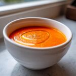 Close-up of a white bowl filled with vibrant Vegan Creamy Tomato Coconut Soup, drizzled with oil and pepper.