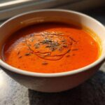 A vibrant bowl of Vegan Creamy Tomato Coconut Soup garnished with oil and herbs.