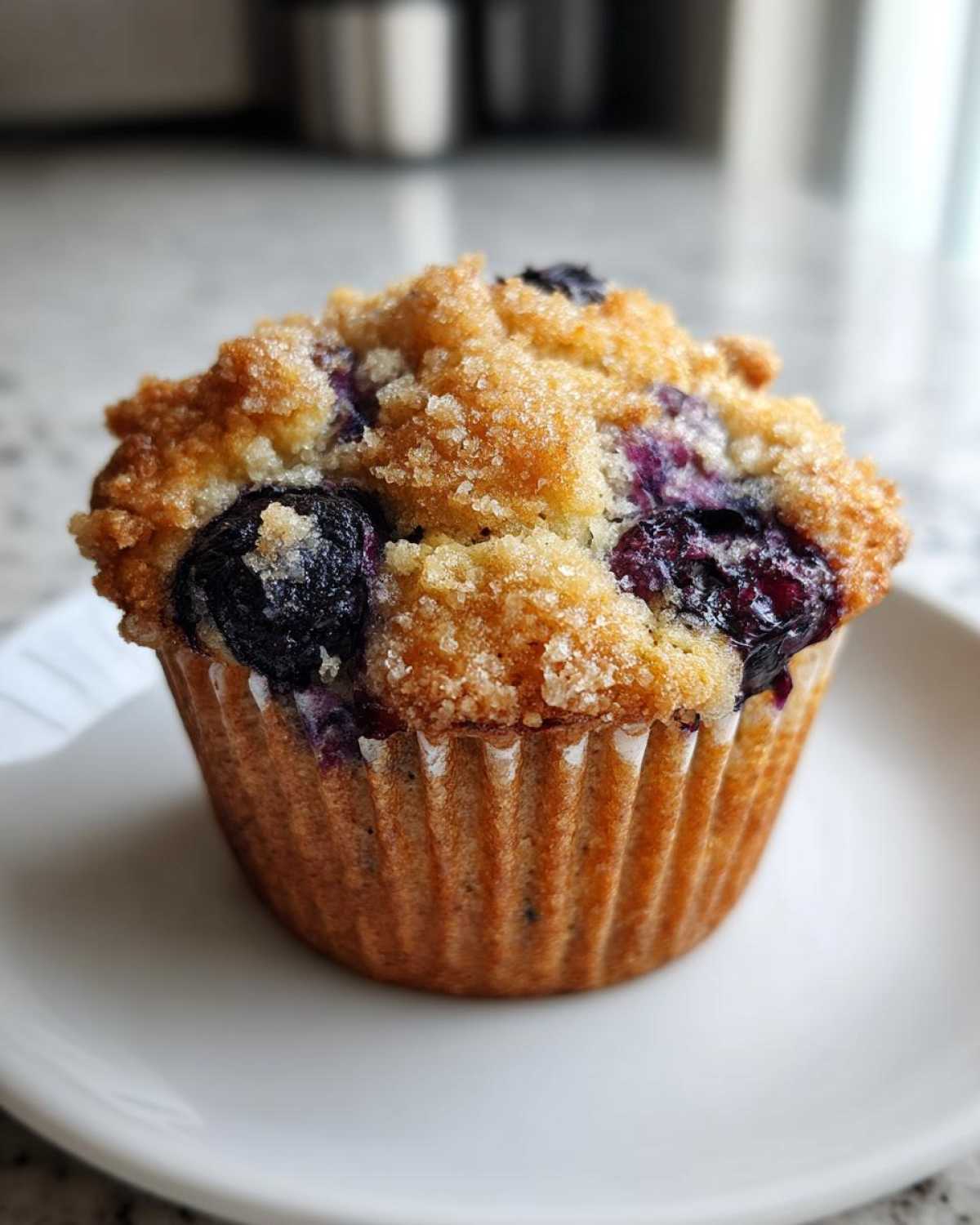 A single, perfectly baked Vegan Lemon Blueberry Muffin topped with coarse sugar, resting on a white plate.
