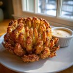A perfectly cooked, golden-brown Air Fryer Blooming Onions served on a white plate next to a small ramekin of dipping sauce.