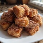 A pile of golden Air Fryer Crispy Cinnamon Sugar Biscuit Bites coated heavily in cinnamon sugar on a white plate.